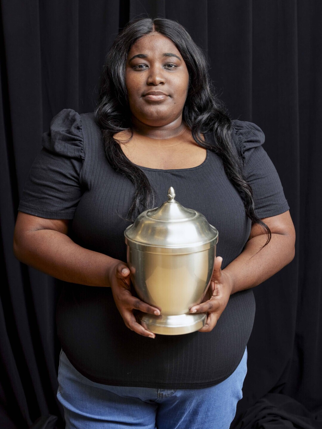A student in all black stands in front a black backdrop looking directly at the camera, holding a large, light gold urn. 