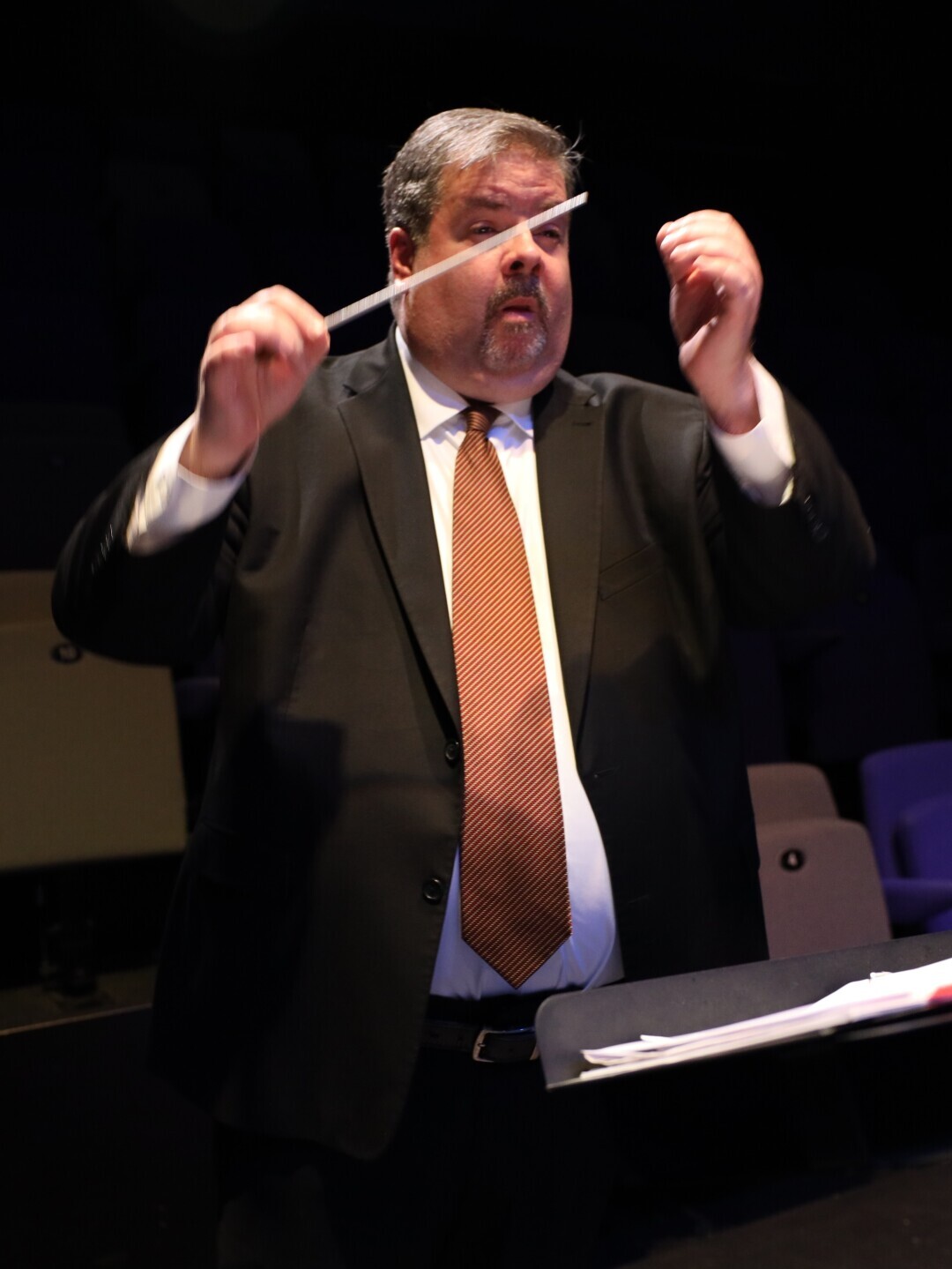 Dr. David Guthrie stands in a darkened theatre in a dark suit and tie, arms raised conducting a musical performance. 