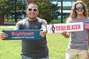 Two students wearing sunglasses and smiling as they stand and hold Brightpoint and Trailblazer signs.