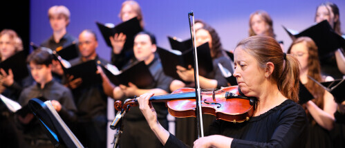 A violinist reads music. A chorus sings behind them.