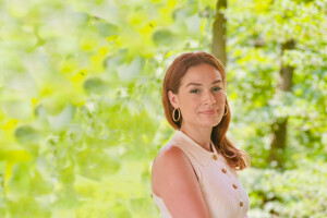 A woman poses with her arm resting on a wooden bar. The background is a bright green forest. 