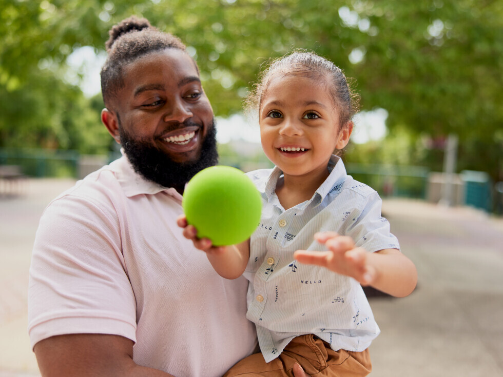 Brightpoint student Jason Taylor holds his son in his arms and smiles at him. His son is also smiling and holding a green ball. 