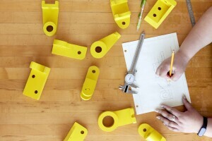 Close up of hands creating technical drawings on paper with a pencil on a light wooden table, surrounded by bright yellow parts and precise measuring tools. 