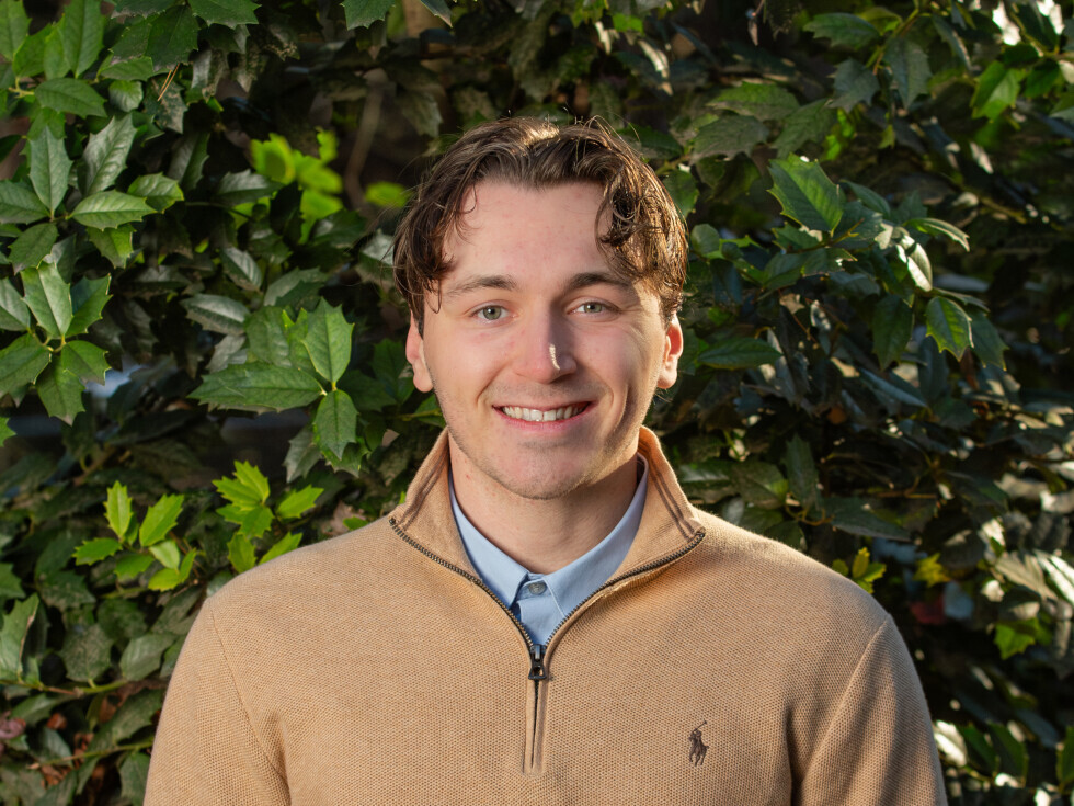 A Brightpoint alum wears a brown sweater and smiles in front of a background of green foliage. 