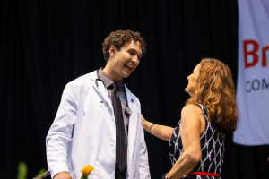 A Spring 2025 nursing graduate wears a white coat and smiles while receiving a pin and yellow rose on stage. 