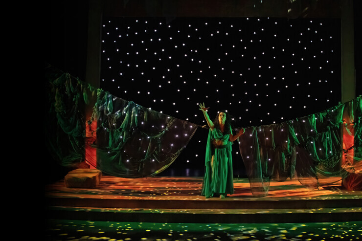 An actor is on a darkened stage in a flowing green dress, surrounded by glowing stars. 