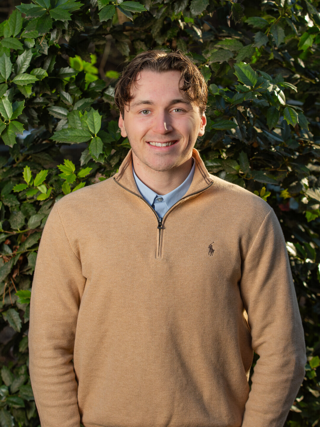 A Brightpoint alum wears a brown sweater and smiles in front of a background of green foliage. 
