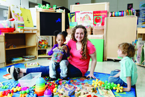 A teacher and two young children sit in an early childhood classroom that includes books, a display board, and creative supplies. 