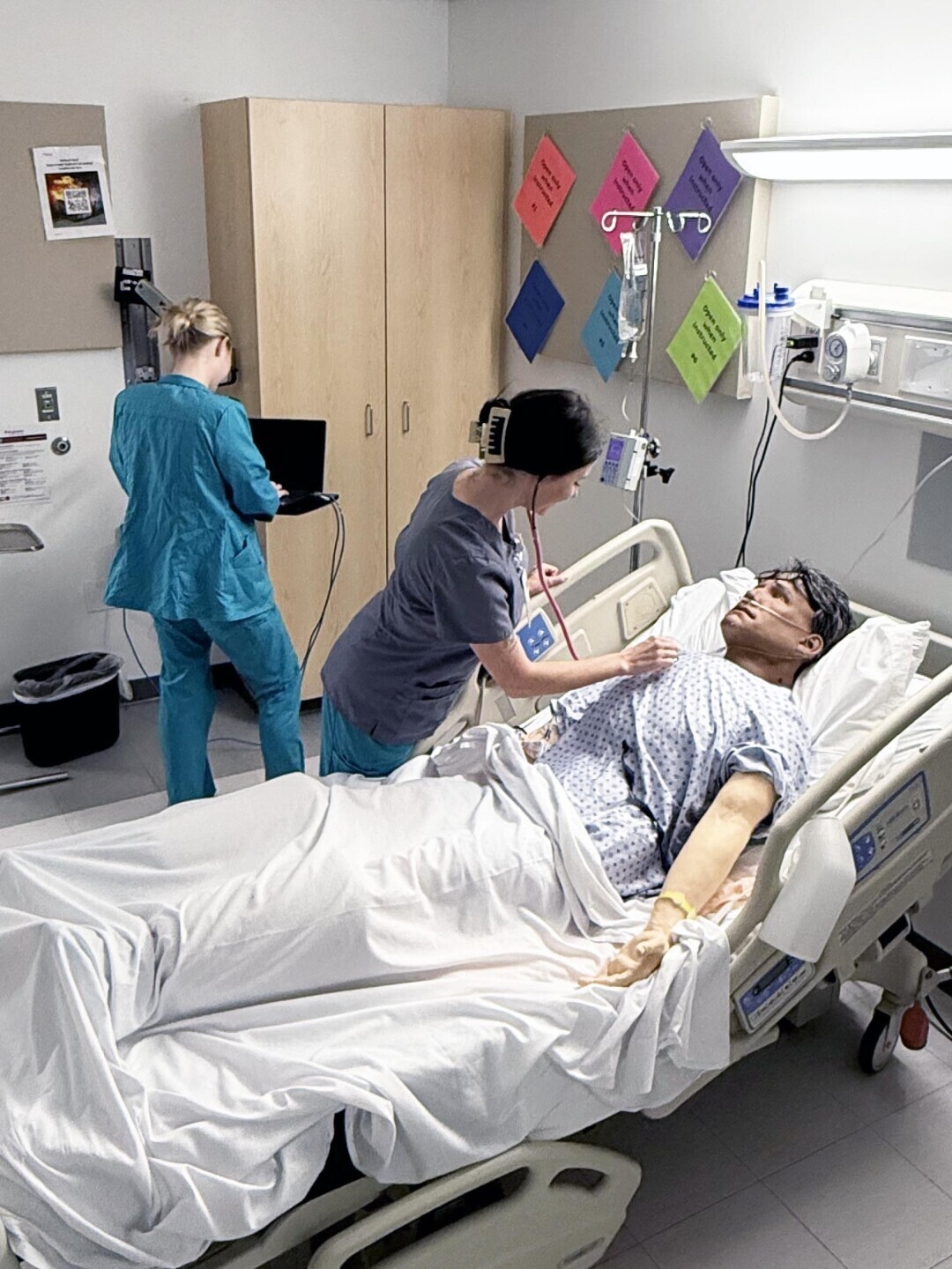 One nursing student is at computer, while another student works on a patient simulator checking vitals on a patient simulator in a hospital bed. 