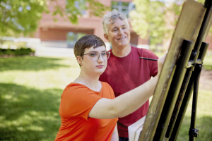 A Brightpoint student paints on an easel, their instructor watching behind them.