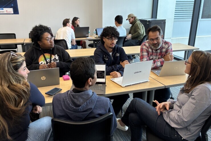 A group of six participants in the AI for Good Hackathon at Brightpoint sit in a tight circle at a table with laptops in front of them engaged in conversation and brainstorming. 