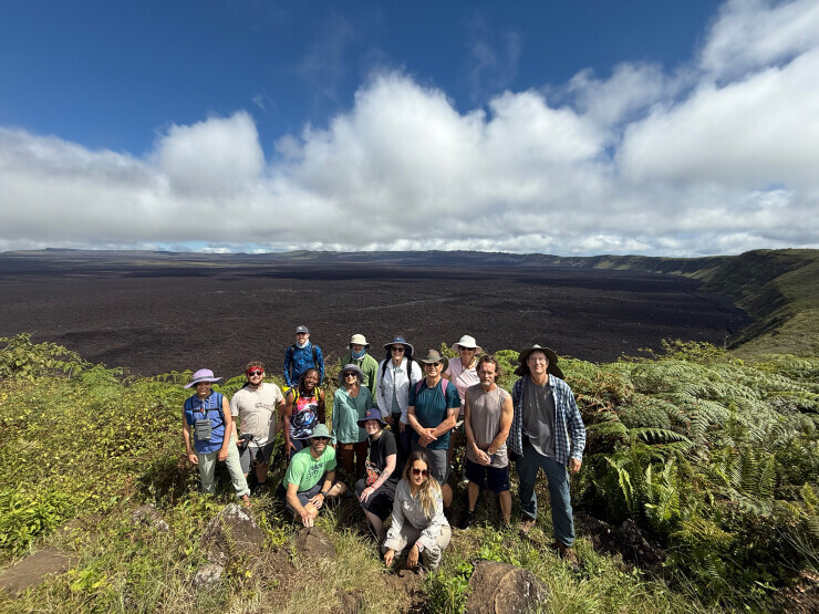 Brightpoint students enjoying a study abroad trip to the Galapagos Islands. The group poses in front of lush vegetation, with a blue sky and volcano behind them. 