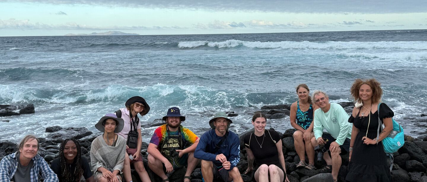 Students on a study abroad trip to the Galapagos Islands. They sit on rocks in front of a blue ocean, smiling.