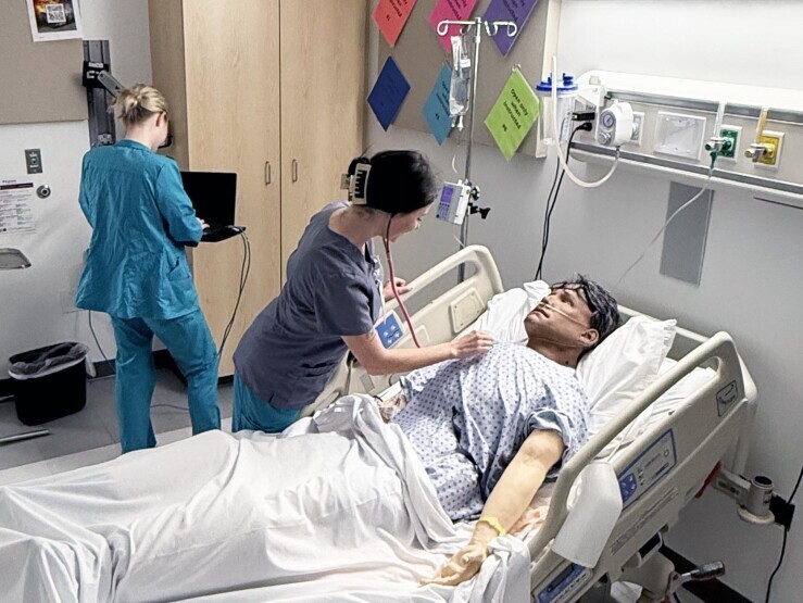 One nursing student is at computer, while another student works on a patient simulator checking vitals on a patient simulator in a hospital bed. 