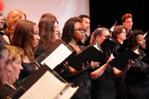 A group of choral performers on stage, all in black, singing and holding folders with sheets of music. 