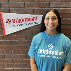 Andrea, a Brightpoint Student Ambassador, smiles in front of a Brightpoint flag. She wears a blue Brightpoint t-shirt.