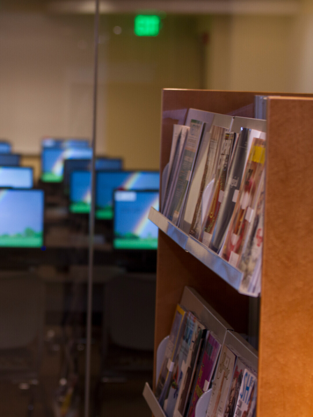 A library bookshelf is full of magazines. Behind it are rows of desktop computers.