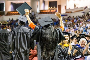Graduate wearing cap and gown raising hands in celebration at Commencement.