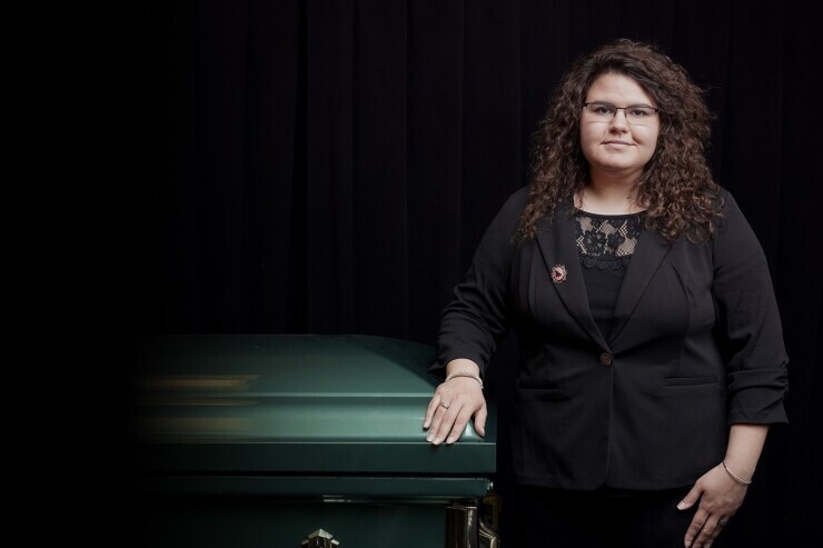 A student dressed in all black professional clothing stands in a darkened room next to a casket with their left hand resting on the top, staring confidentially at the camera. 
