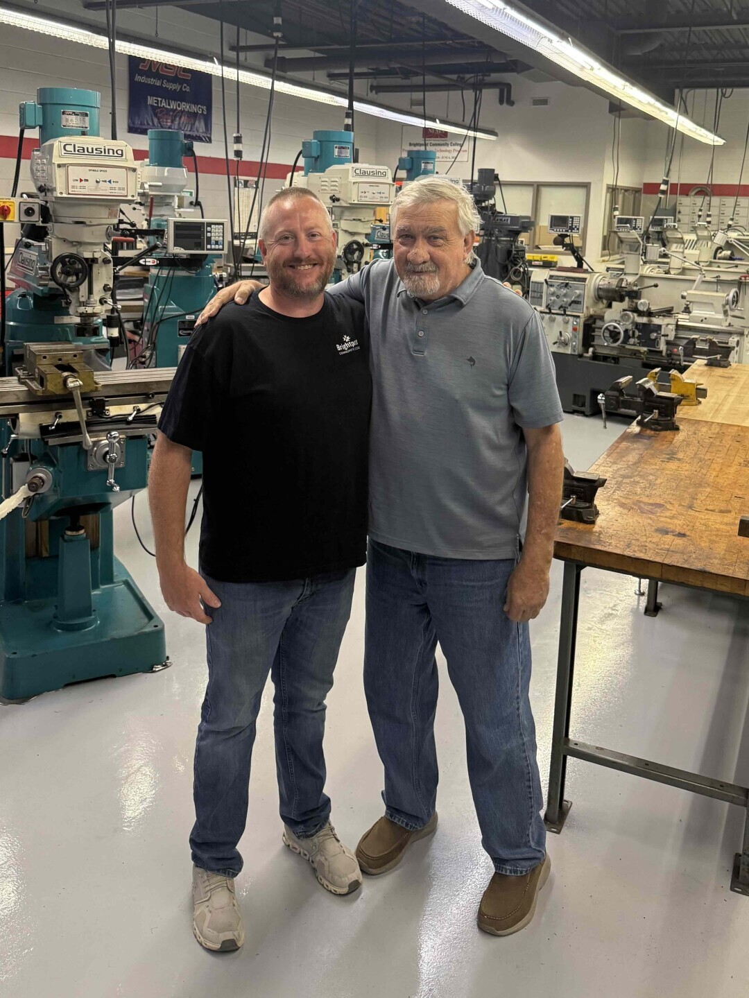 Dustin and Les Bell stand with their arms around each other's shoulders in Brightpoint's precision machining lab surrounded by wooden tables, manual tools and large equipment. 