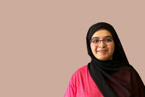 Aneeza Khan smiles softly in front of a pale peach background. 