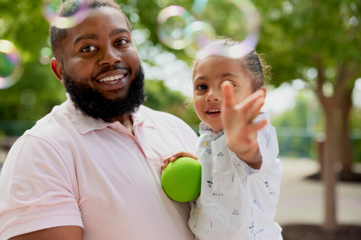 links to A Brightpoint student smiles while holding their young child, who is reaching for a bubble. 
