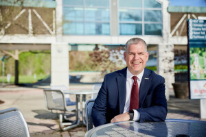 A man in a suit smiling at the camera while seated outdoors. 