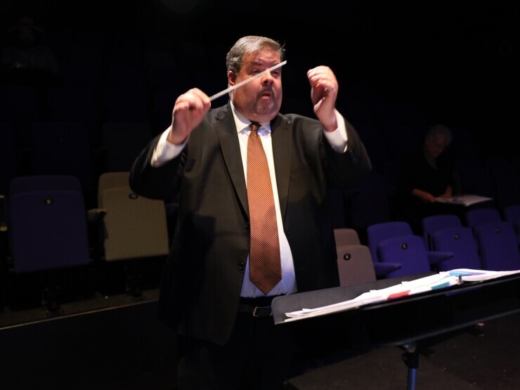 Dr. David Guthrie stands in a darkened theatre in a dark suit and tie, arms raised conducting a musical performance. 