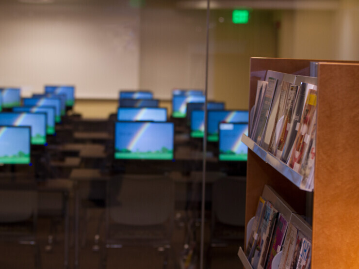A library bookshelf is full of magazines. Behind it are rows of desktop computers.