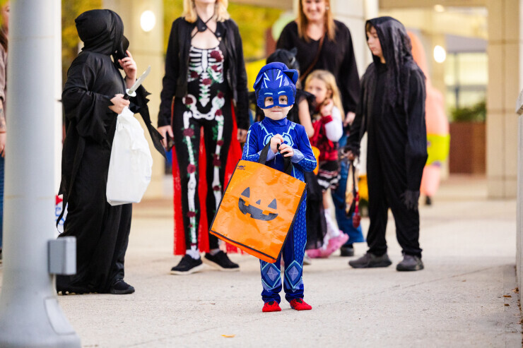 A child wearing a blue superhero costume holds an orange trick-or-treat bag. Behind him are Fall Fest attendees wearing black Halloween costumes. 