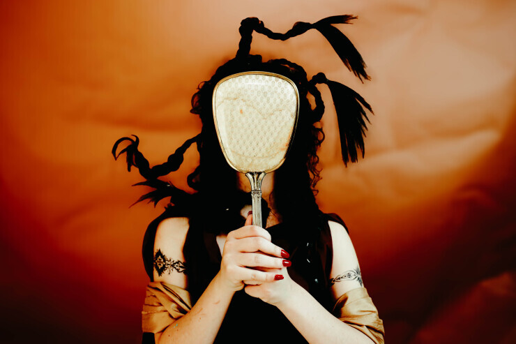 links to A woman holds a mirror to her face, hiding behind it. Her dark hair emerges in braids, and a yellow cloak is draped over her arms. 