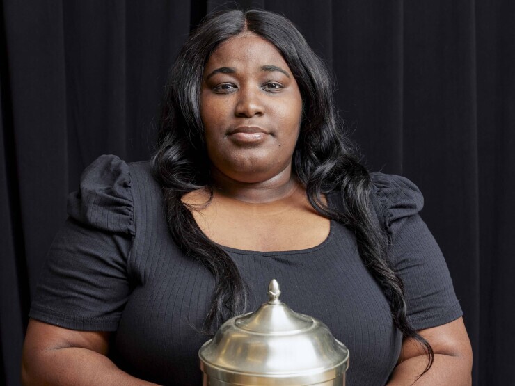 A student in all black stands in front a black backdrop looking directly at the camera, holding a large, light gold urn. 