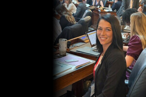 A young woman in a suit smiles while seated at a desk. 
