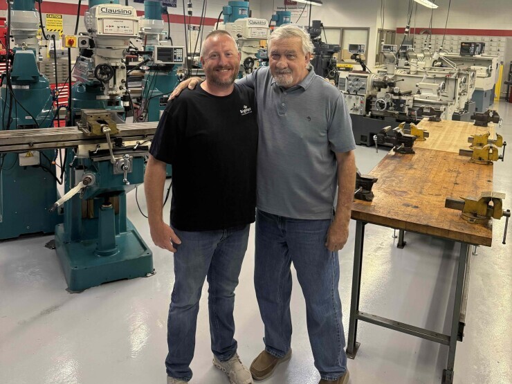 Dustin and Les Bell stand with their arms around each other's shoulders in Brightpoint's precision machining lab surrounded by wooden tables, manual tools and large equipment. 