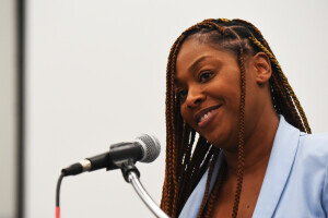 A person with braids smiles while standing in front of a microphone. 