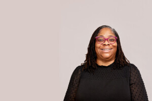 Melinda King wears a black top and poses in front of a taupe background. She smiles. 
